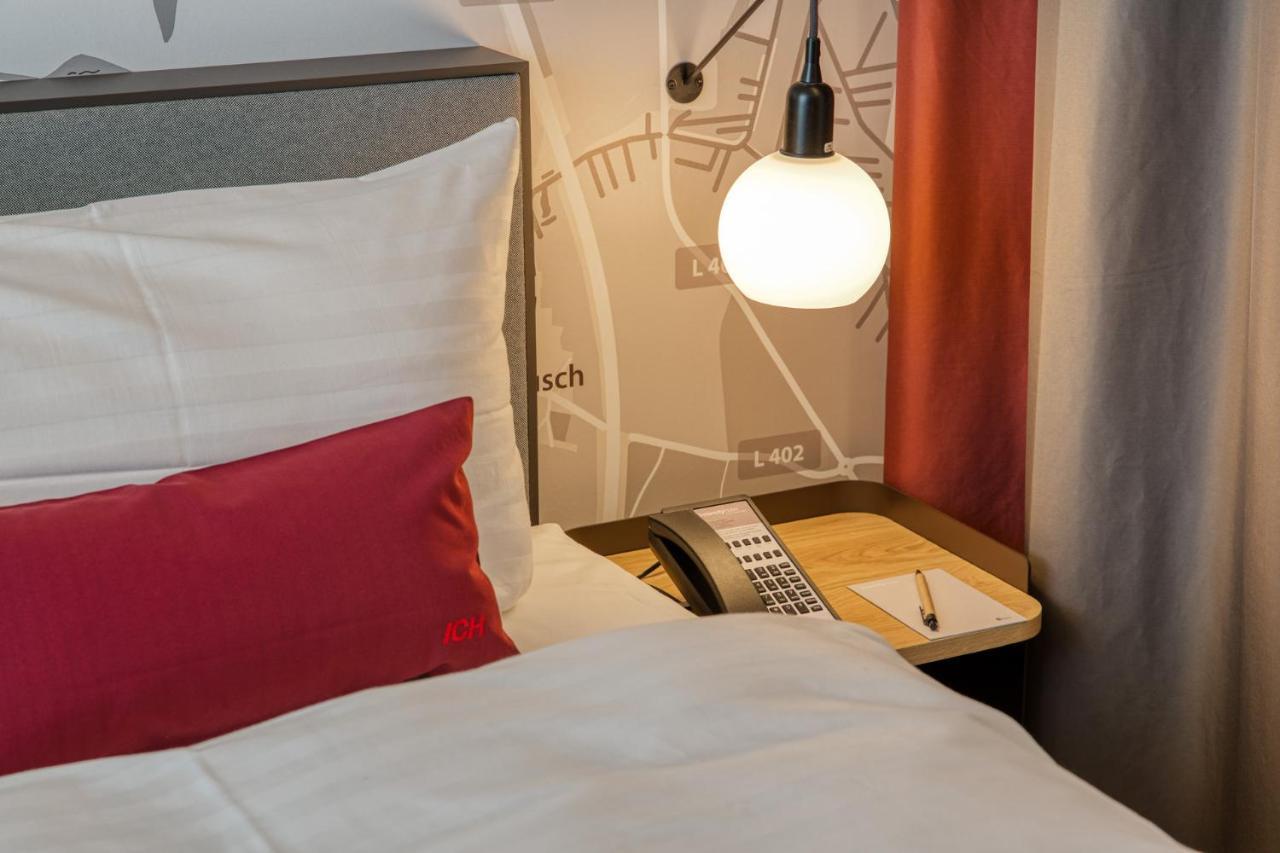 Intercityhotel Berlin Airport Ber Terminal 1 & 2 Schönefeld Exteriör bild The photo shows a cozy bedroom setup. There's a neatly made bed with white bedding and a red decorative pillow. Beside the bed, a small bedside table holds a telephone and a notebook or notepad, along with a pen. A round pendant light hangs above the