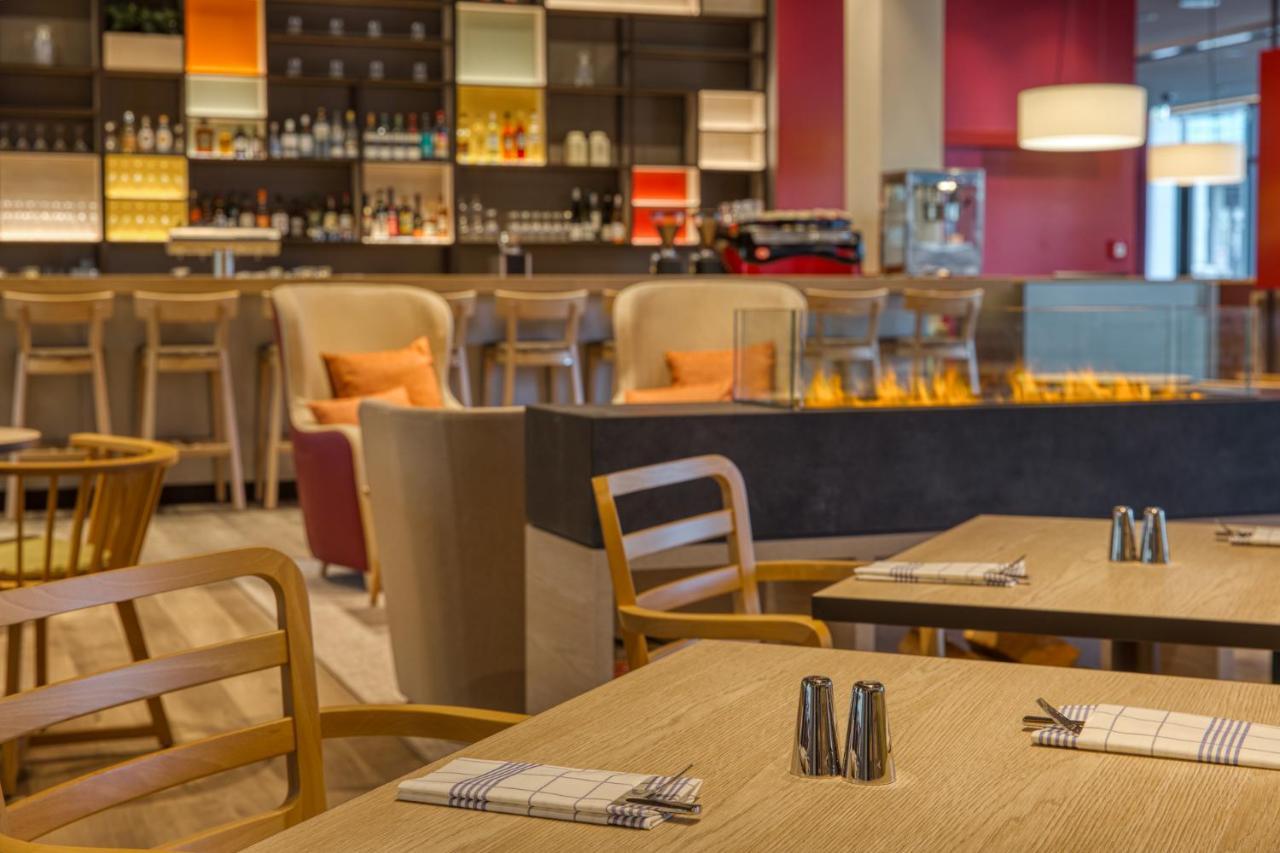 Intercityhotel Berlin Airport Ber Terminal 1 & 2 Schönefeld Exteriör bild The photo shows a modern restaurant or bar interior. In the foreground, there are wooden tables set with napkins and condiments. The seating includes wooden chairs and plush seating in various colors in the background. A stylish bar area is visible w
